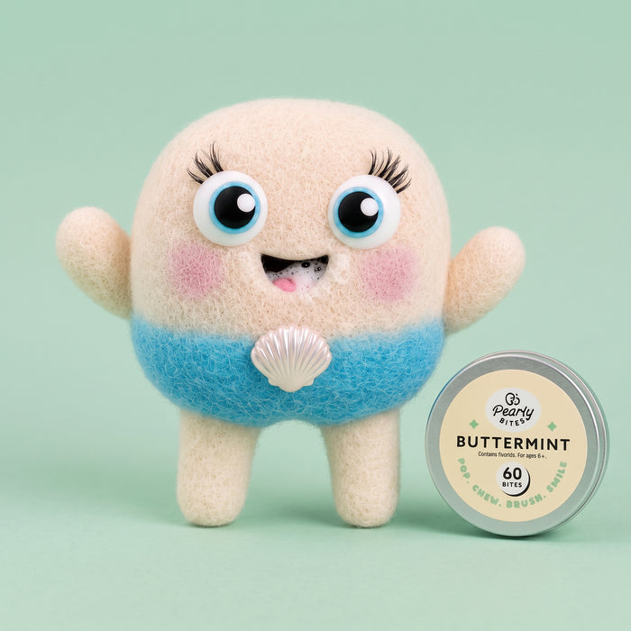A cute, fuzzy mermaid-shaped character toy or plush stands next to a round tin labeled 'Peanut Butter Mint' on a pale mint green background. The character has a cream-colored head and legs, a blue belly, and a white seashell ornament at the center.