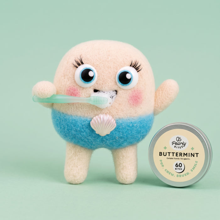 A cute, round, fuzzy character toy with a beige top half, blue bottom half, and a white seashell ornament, holding a mint-green toothbrush. It is shown next to a round tin of Peony Bunnies Buttermint flavored dental chew on a solid mint green background.