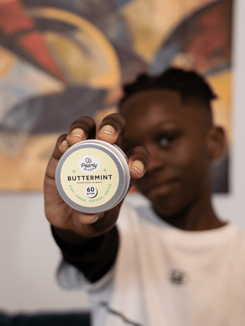 A person holds up a small round tin of Poppy Seed Co. Buttermint Pop Cream Balm toward the camera, with a blurred background and the person's face out of focus.