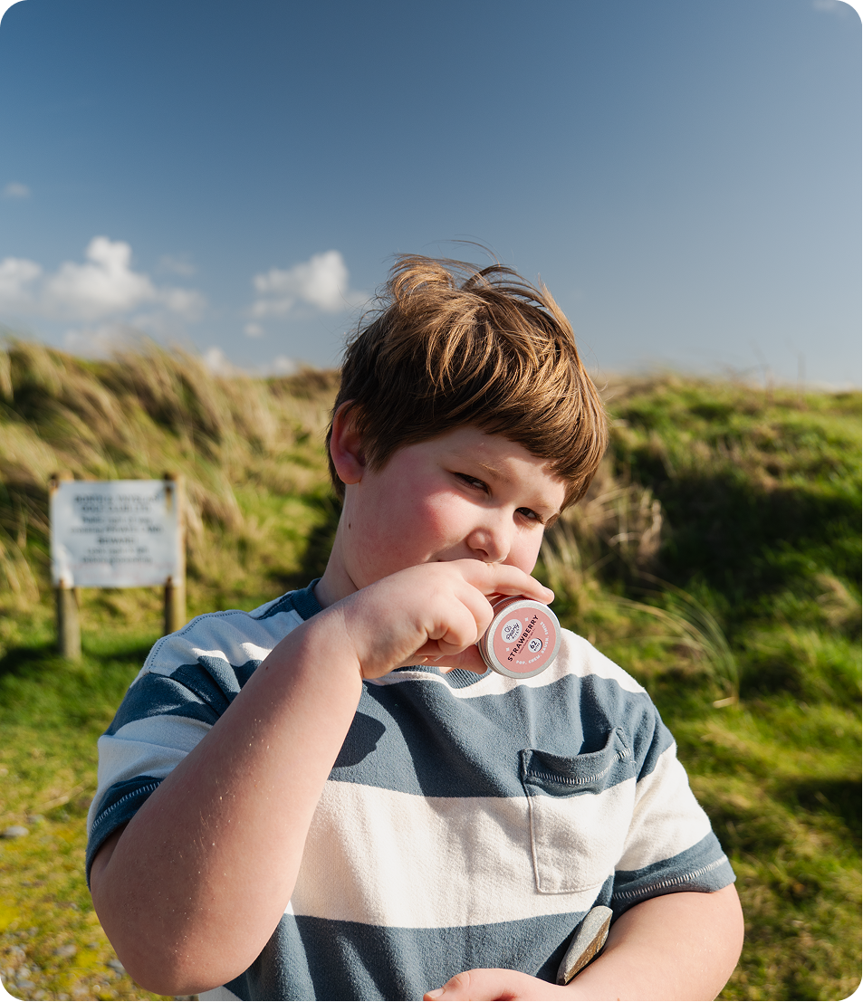 A young boy with brown hair stands outdoors on a grassy hill under a blue sky, wearing a blue and white striped t-shirt and holding a small round tin near his mouth.