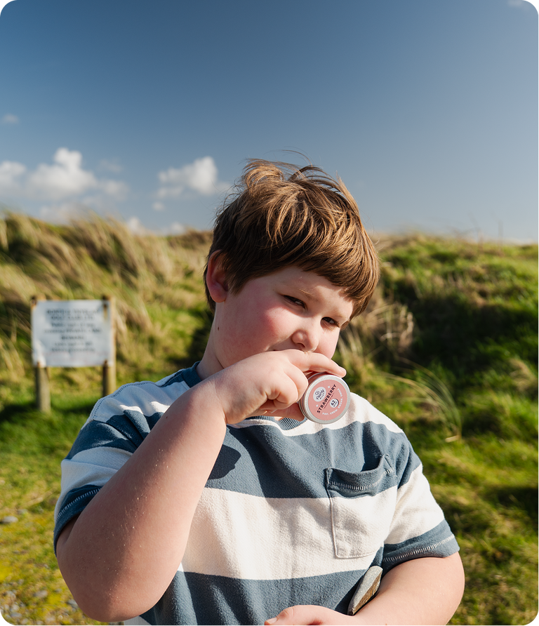A young boy with brown hair stands outdoors on a grassy hill under a blue sky, wearing a blue and white striped t-shirt and holding a small round tin near his mouth.