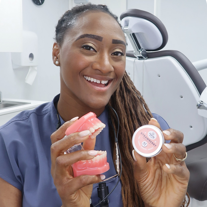 A smiling dental professional in blue scrubs holds a pink dental mold and a small round tin labeled 'strawberry' in a bright dental office setting.