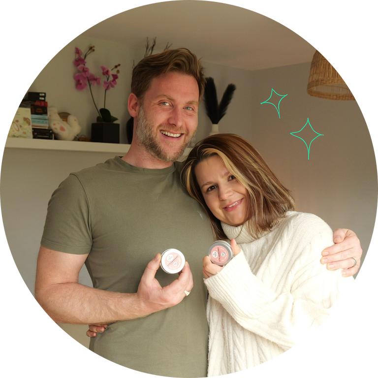 A smiling man and woman standing indoors and holding small round pear;y bites tooth tablets containers. The man wears an olive green T-shirt, and the woman wears a cream cable-knit sweater