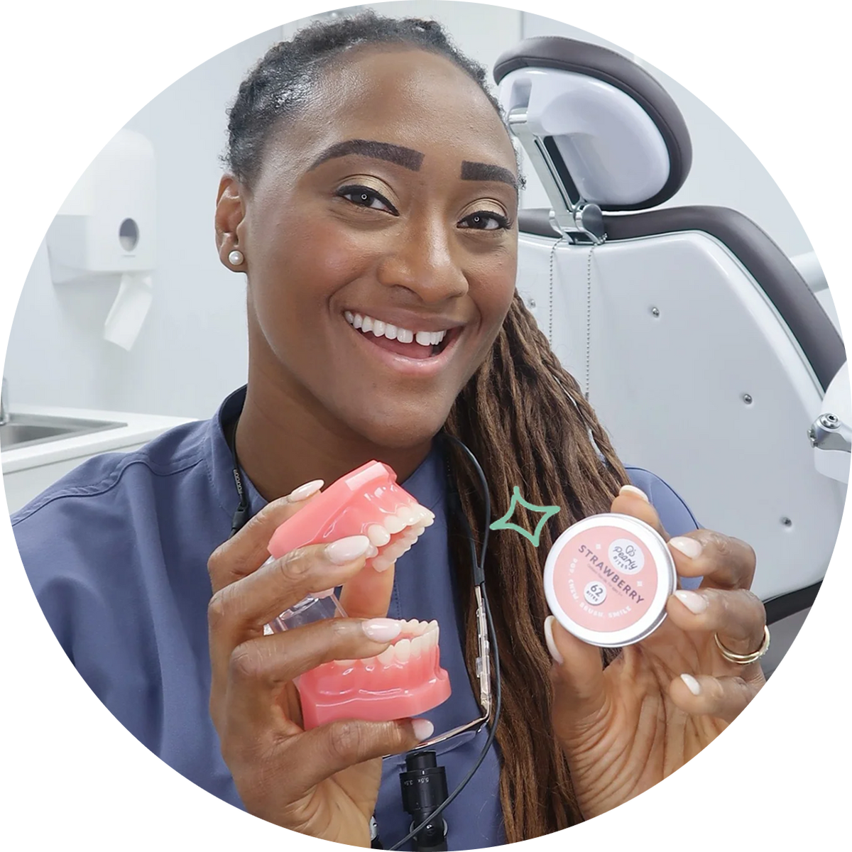 A smiling dental professional in blue scrubs holds a pink dental model and a small round container labeled 'STRAWBERRY' in a bright dental office setting.