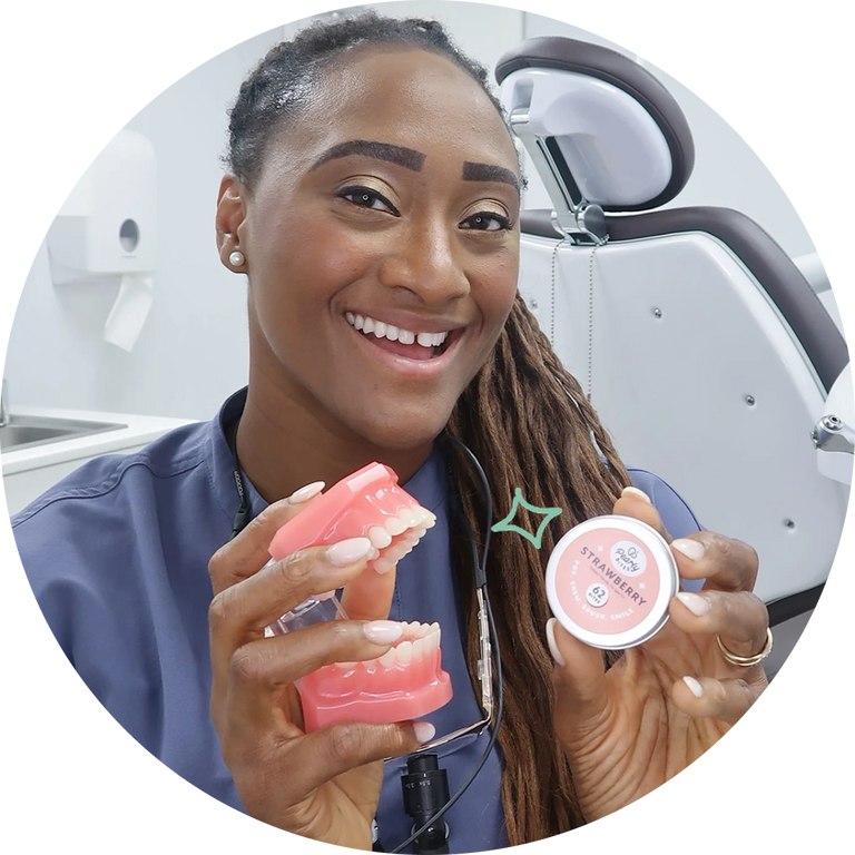 A smiling dental professional in blue scrubs holds a pink dental model and a small round container labeled 'STRAWBERRY' in a bright dental office setting.