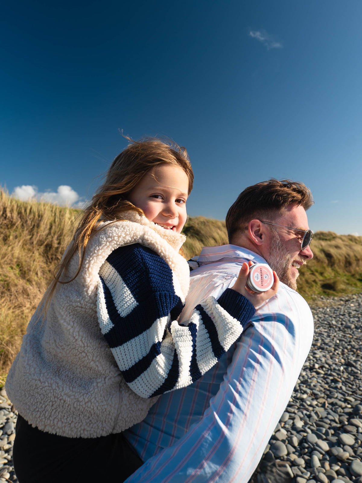 A man carrying a young girl on his back outdoors on a pebble beach, with dune grass and a clear blue sky in the background. The girl wears a cream sherpa vest over a navy and white striped sweater, and the man wears a light blue striped shirt.