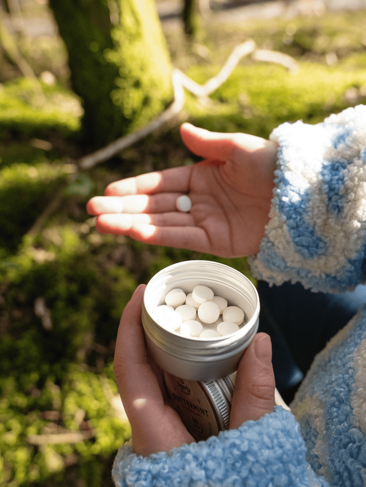 A person outdoors holds an open silver tin filled with small white tablets, with one tablet resting on their palm. The background shows a blurred natural setting with green moss and tree trunks.