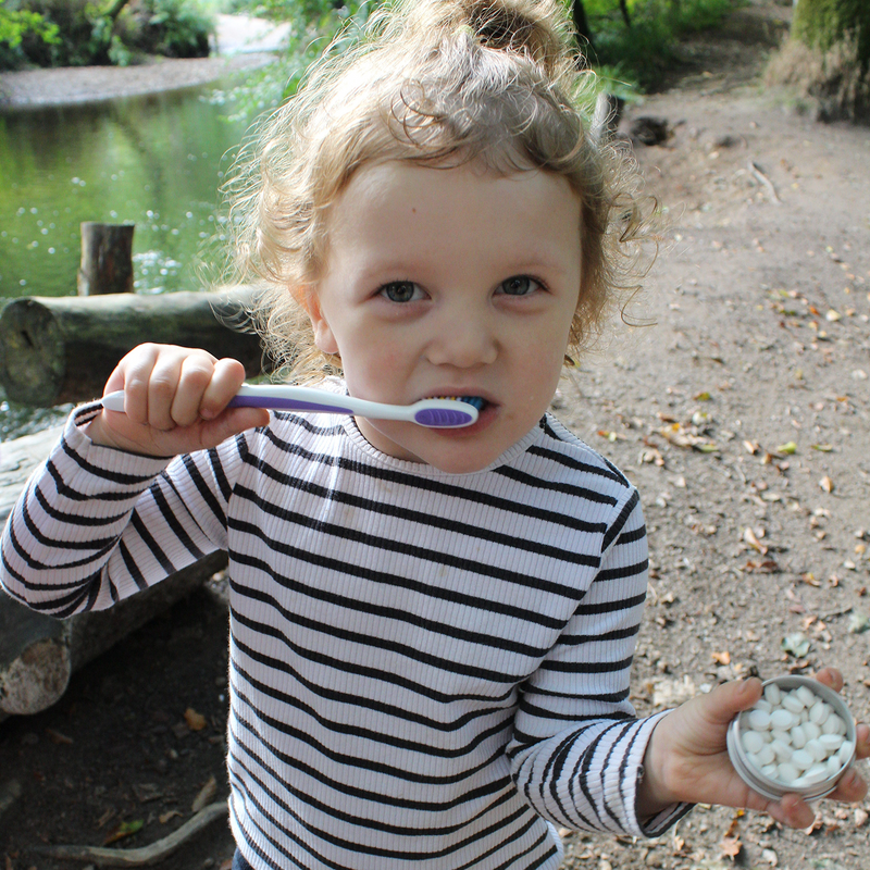 A young child with curly blonde hair wearing a black and white striped shirt, brushing their teeth with a purple and white toothbrush while holding a small tin of white dental floss in an outdoor park setting.
