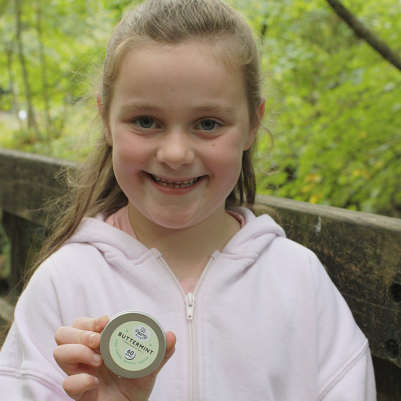 A young girl with light brown hair smiles while holding a small round tin of Bittermint tea outdoors. She is wearing a light pink zip-up hoodie, with a wooden railing and green foliage blurred in the background.