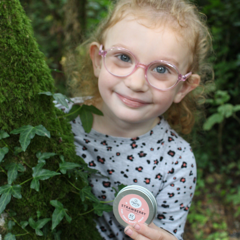 A young girl with curly blonde hair and pink glasses smiles while holding a small round tin of strawberry pearly bites tooth tablets outdoors among green moss and ivy.