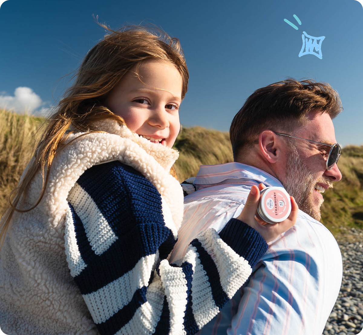 A smiling woman carrying a child on her back outdoors under a blue sky. The child wears a cream sherpa-lined striped sweater, and the woman holds a small round product tin